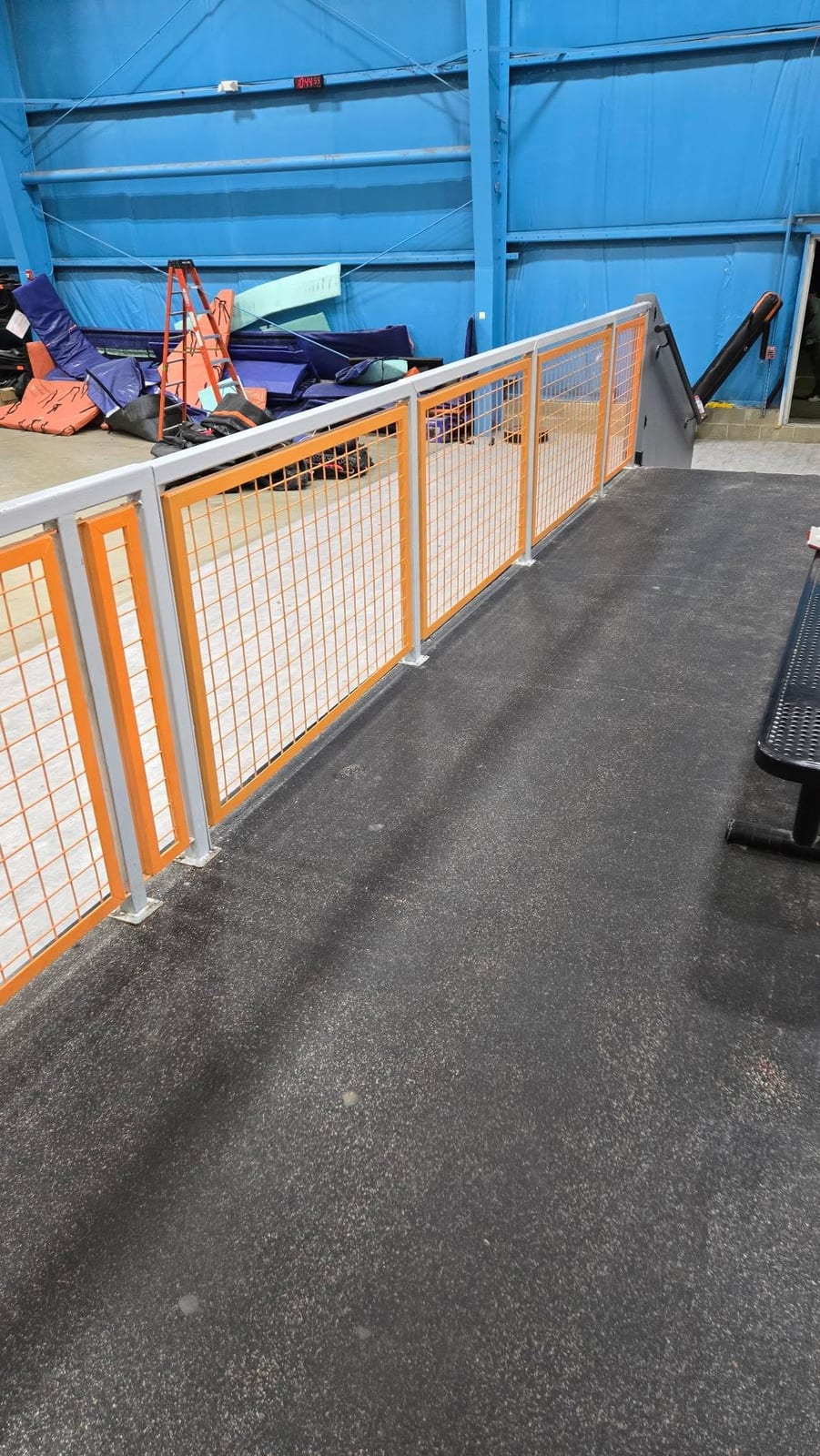 A gray and orange steel guardrail with welded wire mesh panels installed along the floor of an indoor recreation facility