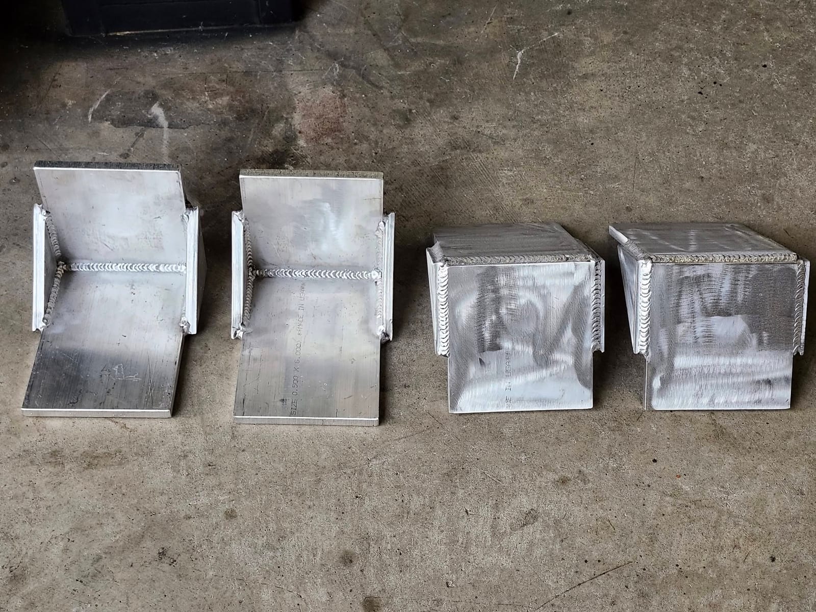 Four heavy half inch plate brackets with evenly rippled weld beads lined up on the shop floor