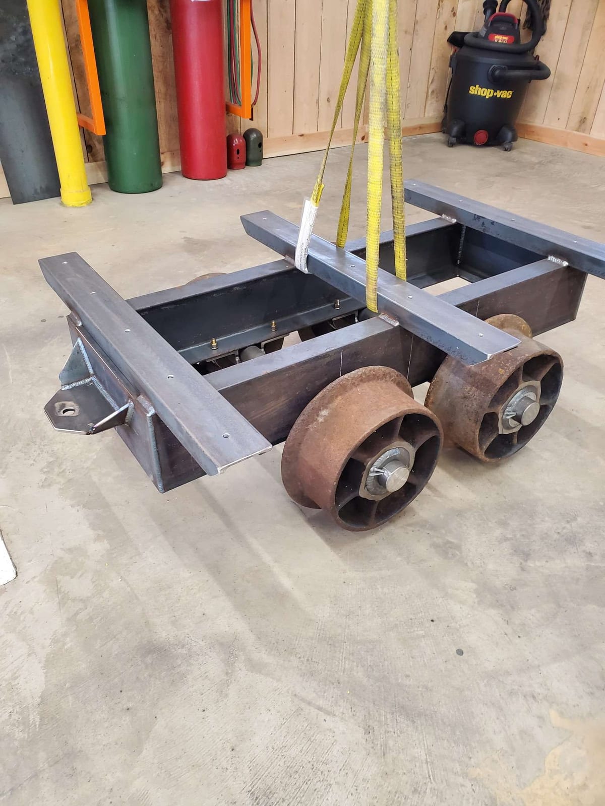 A fabricated steel rail cart with a channel frame, angle iron side rails and four cast iron rail wheels, hanging from a lifting strap on the Robinson Welding shop floor