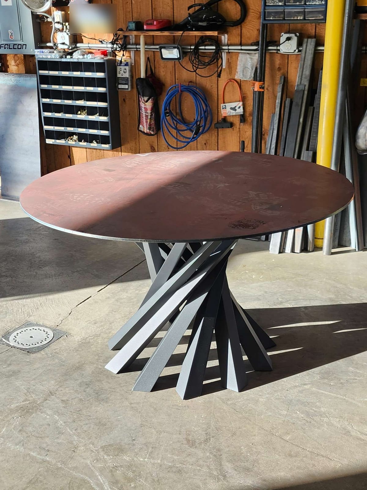 A round steel plate table top on a pedestal base made of fanned and twisted steel bars, standing in the Robinson Welding shop