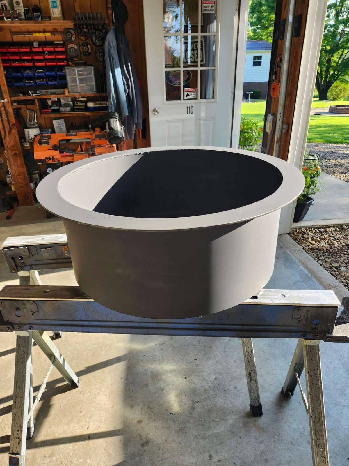 A rolled steel fire pit ring insert with a wide flat top flange, freshly primed, sitting on sawhorses at the open door of the Robinson Welding shop