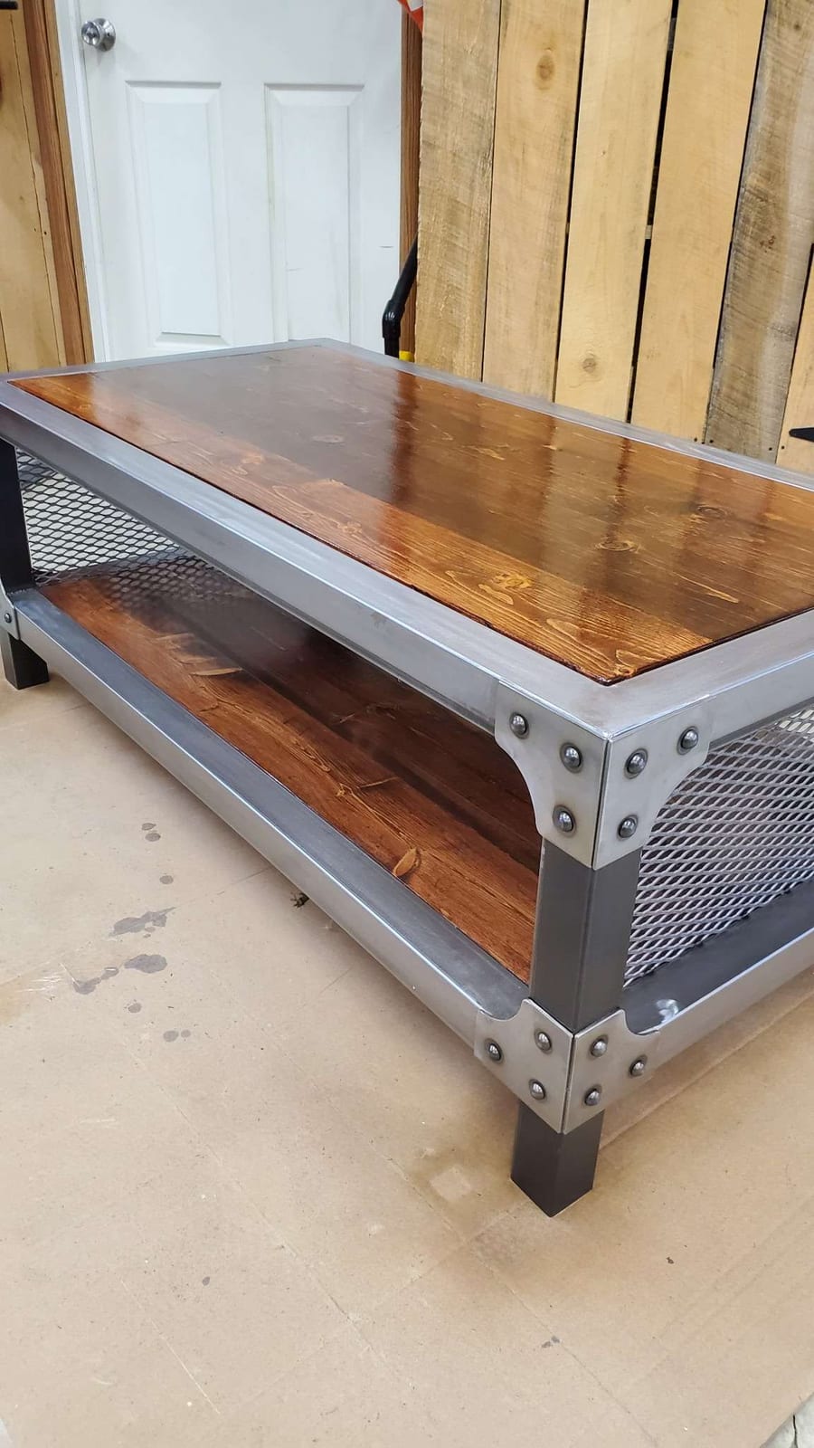 A custom coffee table with a polished steel angle frame, riveted corner gussets, expanded metal end panels, a stained wood top and a matching wood lower shelf