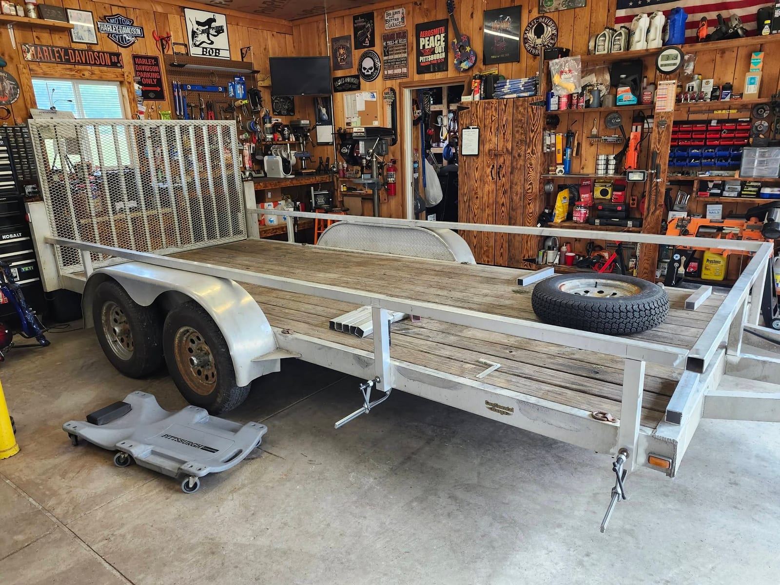 An aluminum utility trailer with a wood deck and mesh ramp gate parked inside the shop for frame repair