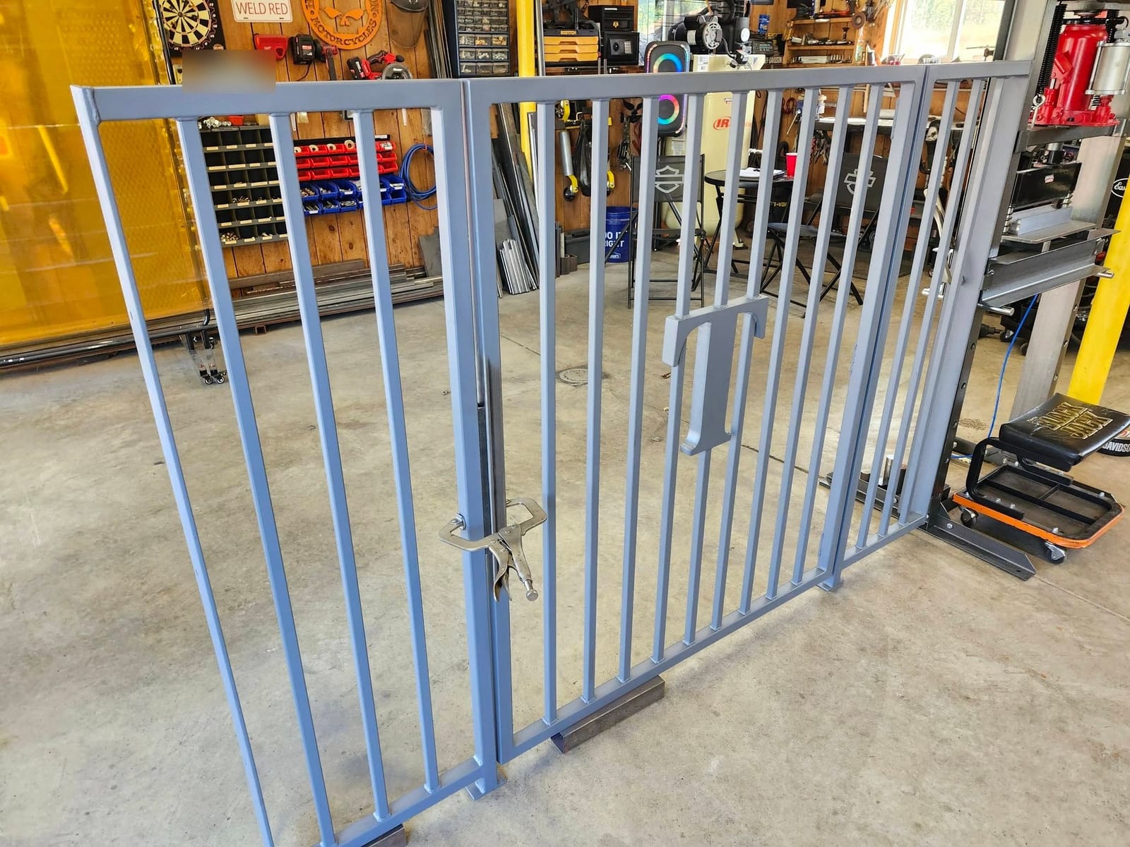 A custom steel name gate painted gray, with a cut plate letter T set between the spindles, standing in the Robinson Welding shop